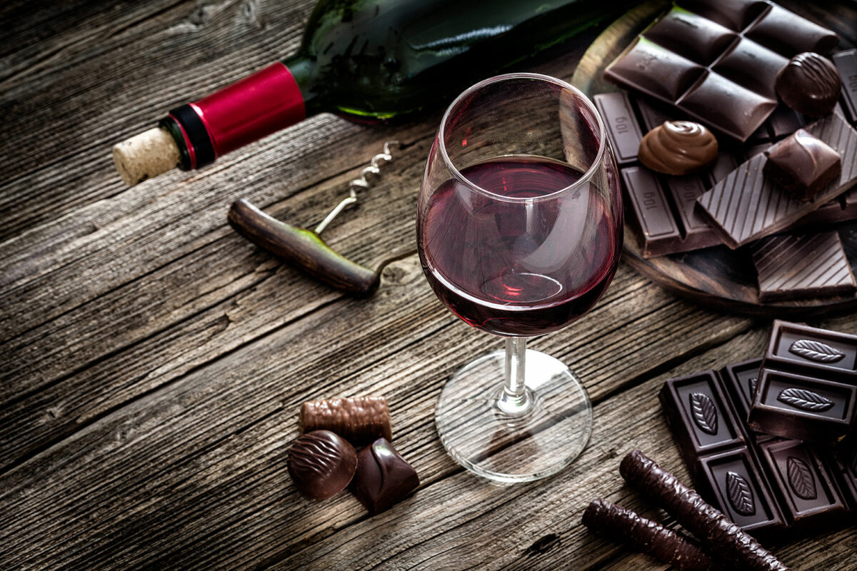 Ein Tisch, auf dem ein gefülltes Rotweinglas steht und eine Rotweinflasche samt Korkenzieher liegen - daneben liegen Schokoladentafeln und Pralinen.