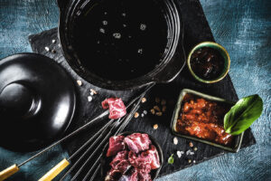 Fondue-Topf mit heißem Fett auf einer Schieferplatte, daneben Spieße mit Rindfleisch und einem Dip.