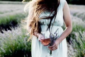 Frau steht in einem Lavendelfeld und hält einen Lavendelstrauß sowie ein Glas mit Rosé aus der Provence in den Händen