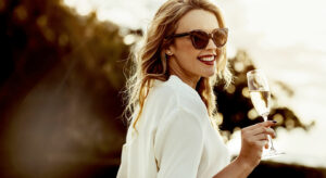 Frau steht mit Sonnenbrille im Sonnenuntergang und hält ein Sektglas mit Perlwein in der Hand